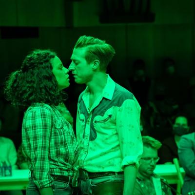Anoushka Lucas as Laurey Williams and Arthur Darvill as Curly McLain standing under green light, facing each other and standing so close together that their faces are almost touching. 