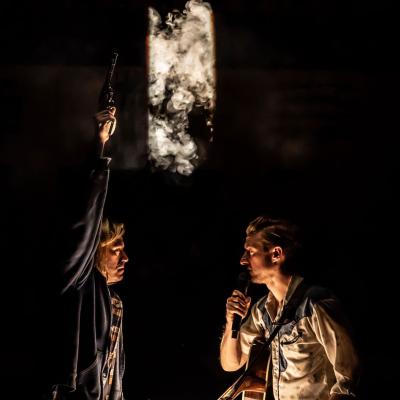 Patrick Vaill as Jud Fry and Arthur Darvill as Curly McLain sat opposite each other in low light, both holding microphones with Patrick Vaill as Jud Fry holding a gun up in the air