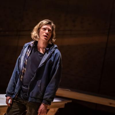 Patrick Vaill as Jud Fry wearing a dark blue hoodie and t-shirt, with parts of a wooden seat in the background. 