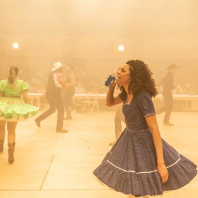 The cast of of Rodgers & Hammerstein's Oklahoma! all dancing in haze with golden light, Anoushka Lucas as Laurey Williams knocks back a can of Beer, in her party dress