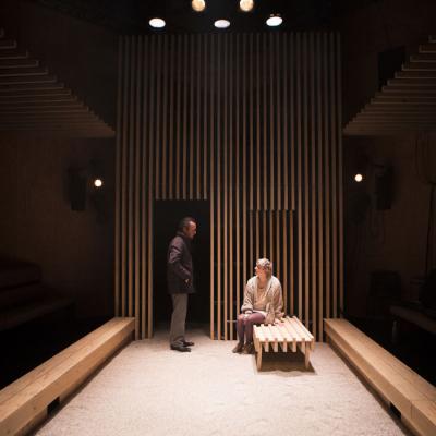 A wide angle shot looking down the traverse stage towards Jonathan Cullen (Man) who is wearing a suit almost standing over Maisie Greenwood (Woman) who is sitting on a bench in Winter at the Young Vic © Ellie Kurttz