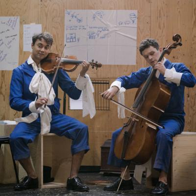 Two male actors sit in their blue jumpsuits with white collars. One is playing a violin, the other a cello © Leon Puplett