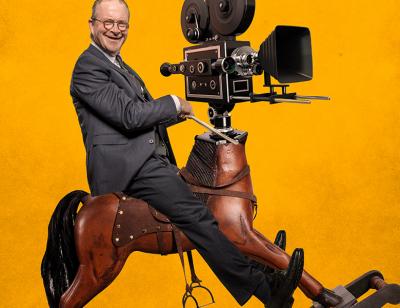 Harry Enfield riding a rocking horse with a old school film camera for a head